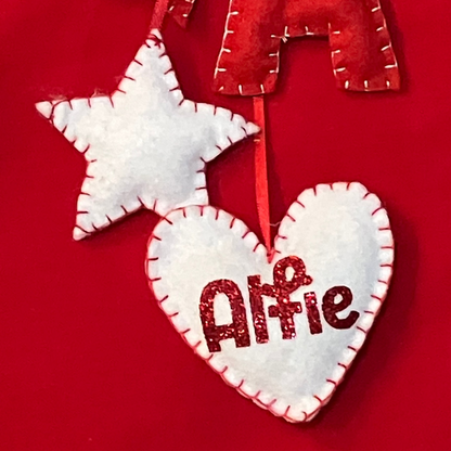 A white felt star and heart, with “Alfie” written on the heart, hanging from a red stocking.