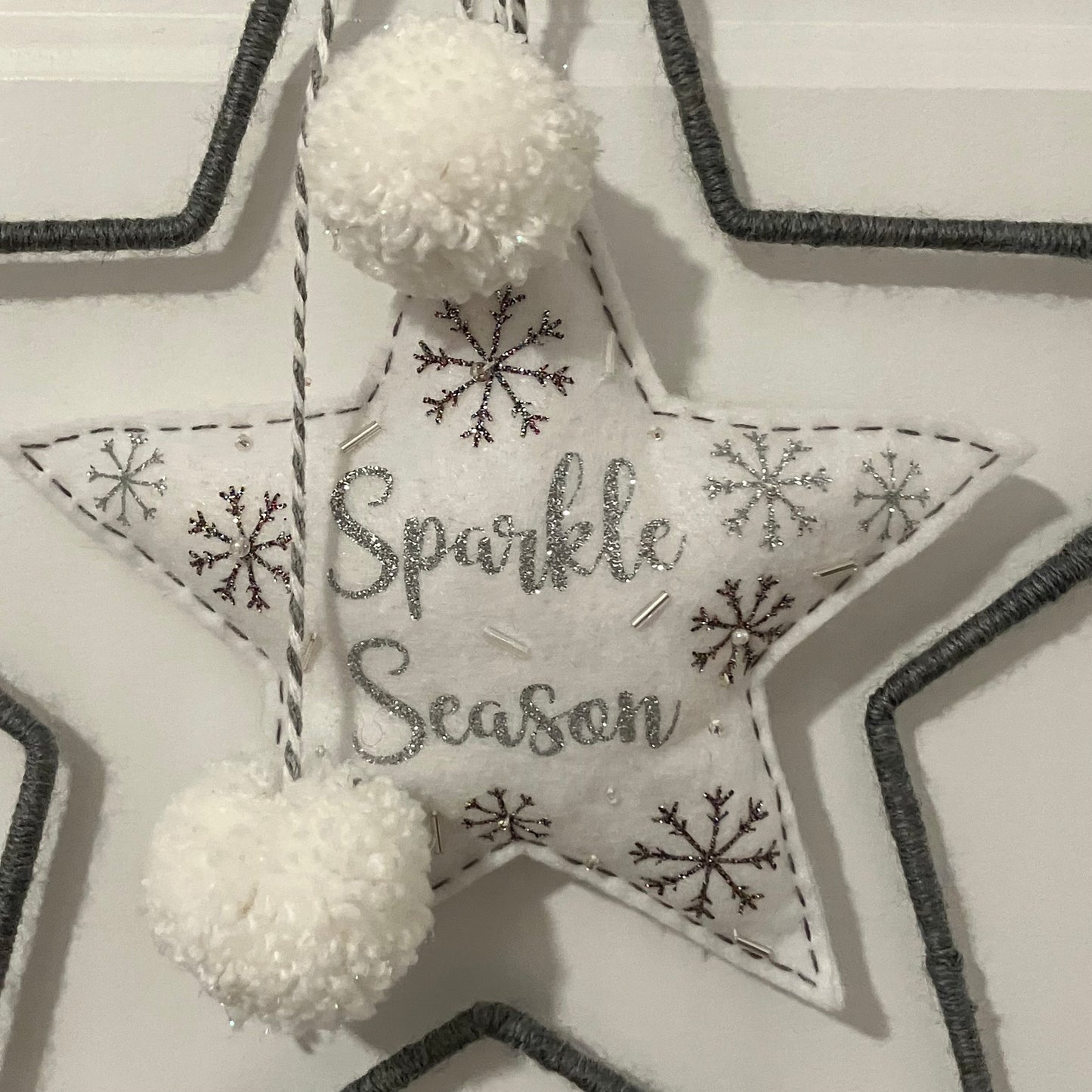 White felt star ornament with “Sparkle Season” embroidered in silver, surrounded by snowflakes and two white pom-poms.