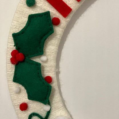 Close-up of a white felt wreath with green holly leaves and red berries.