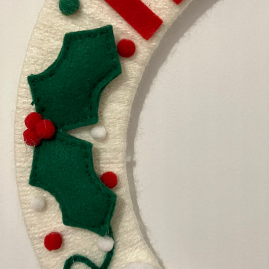 Close-up of a white felt wreath with green holly leaves and red berries.