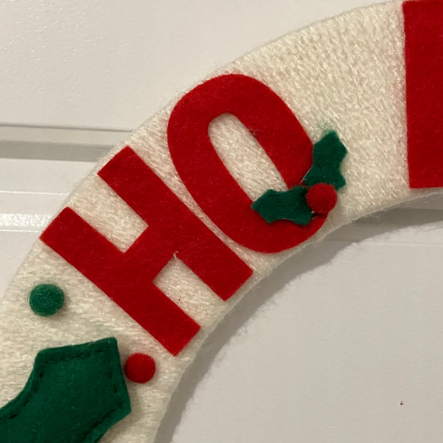 Close-up of a white felt wreath with red letters "HO" and green holly leaves with red berries.