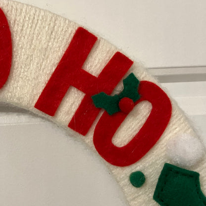 Close-up of a white wreath with red felt letters spelling "HO" and a small holly decoration.
