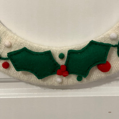 Close-up of a knitted cream-colored wreath with green felt holly leaves and red berries.