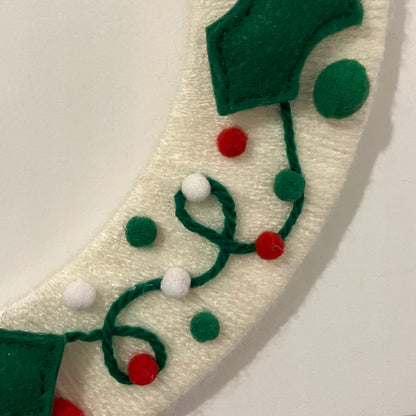 Close-up of a white yarn wreath with green felt leaves and red, white, and green pom-poms.