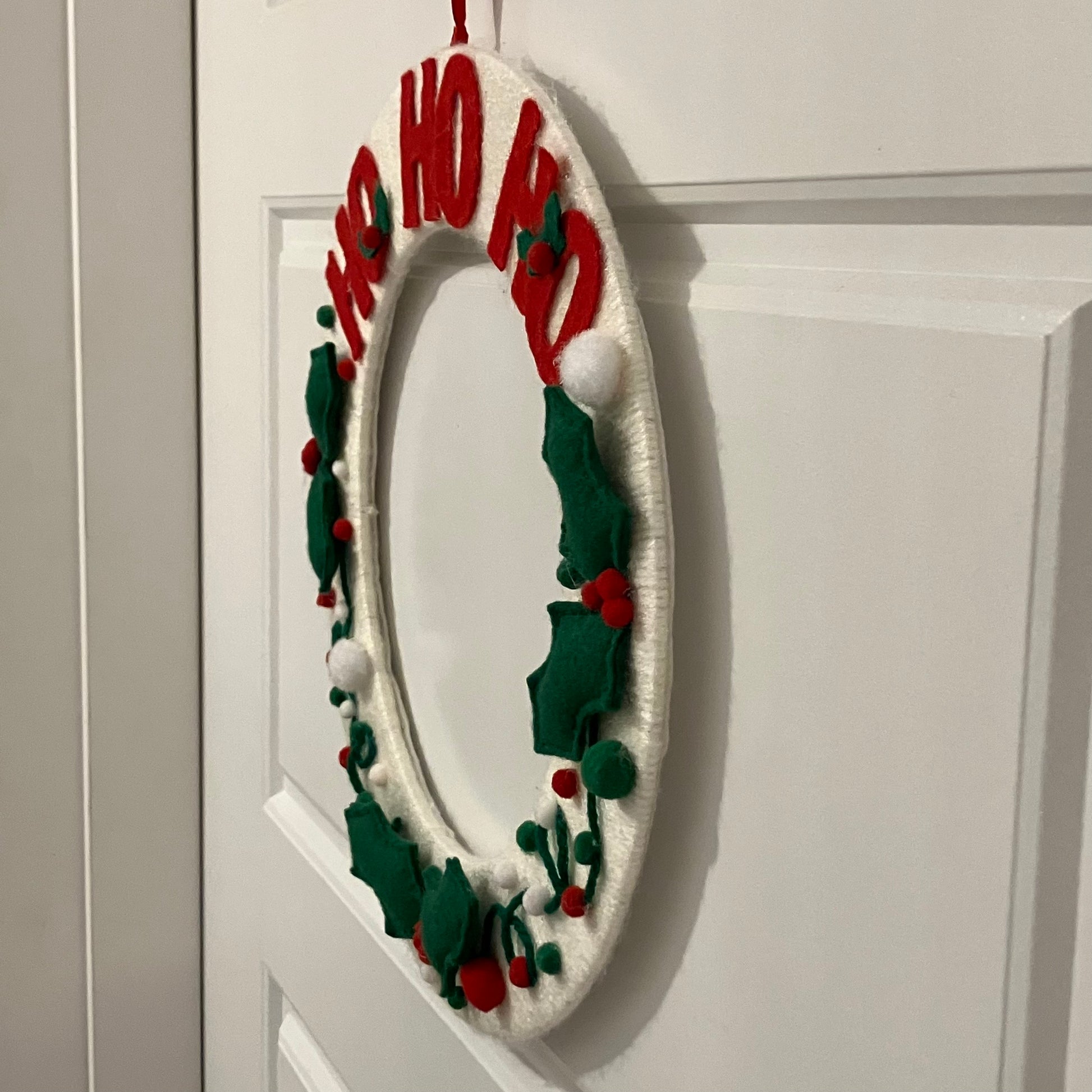 White oval wreath with red "HO HO HO" letters, green holly leaves, and red berries.