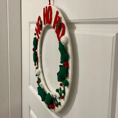 White oval wreath with red "HO HO HO" letters, green holly leaves, and red berries.