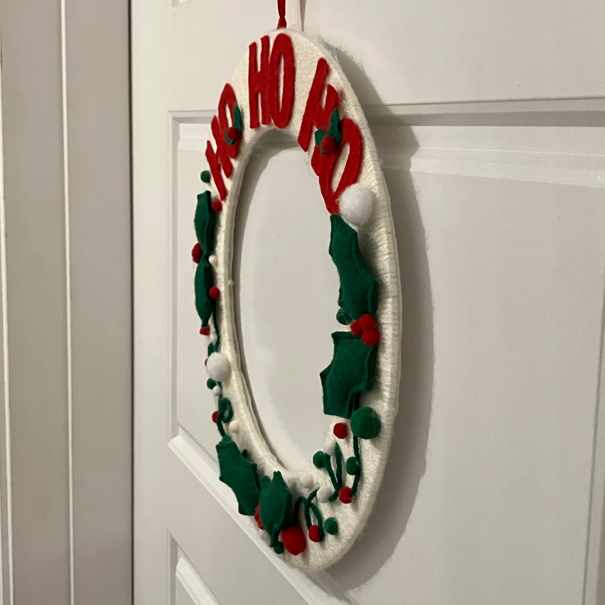 White oval wreath with red "HO HO HO" lettering, green holly leaves, and red berries.