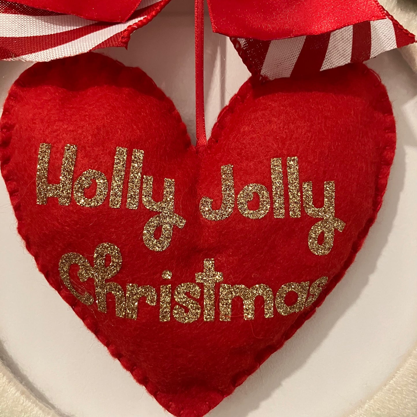 Red felt heart ornament with "Holly Jolly Christmas" in gold glitter, hanging from a red and white striped ribbon.