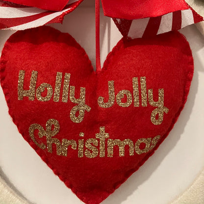 Red felt heart ornament with "Holly Jolly Christmas" in gold glitter, hanging from a red and white striped ribbon.