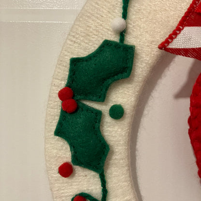 Close-up of a white knitted wreath with green felt holly leaves and red berries.