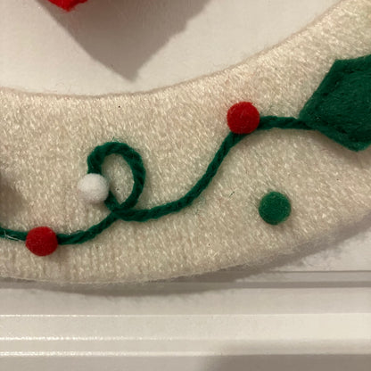 Close-up of a white felt Christmas wreath with green felt garland and red and white pom-poms.
