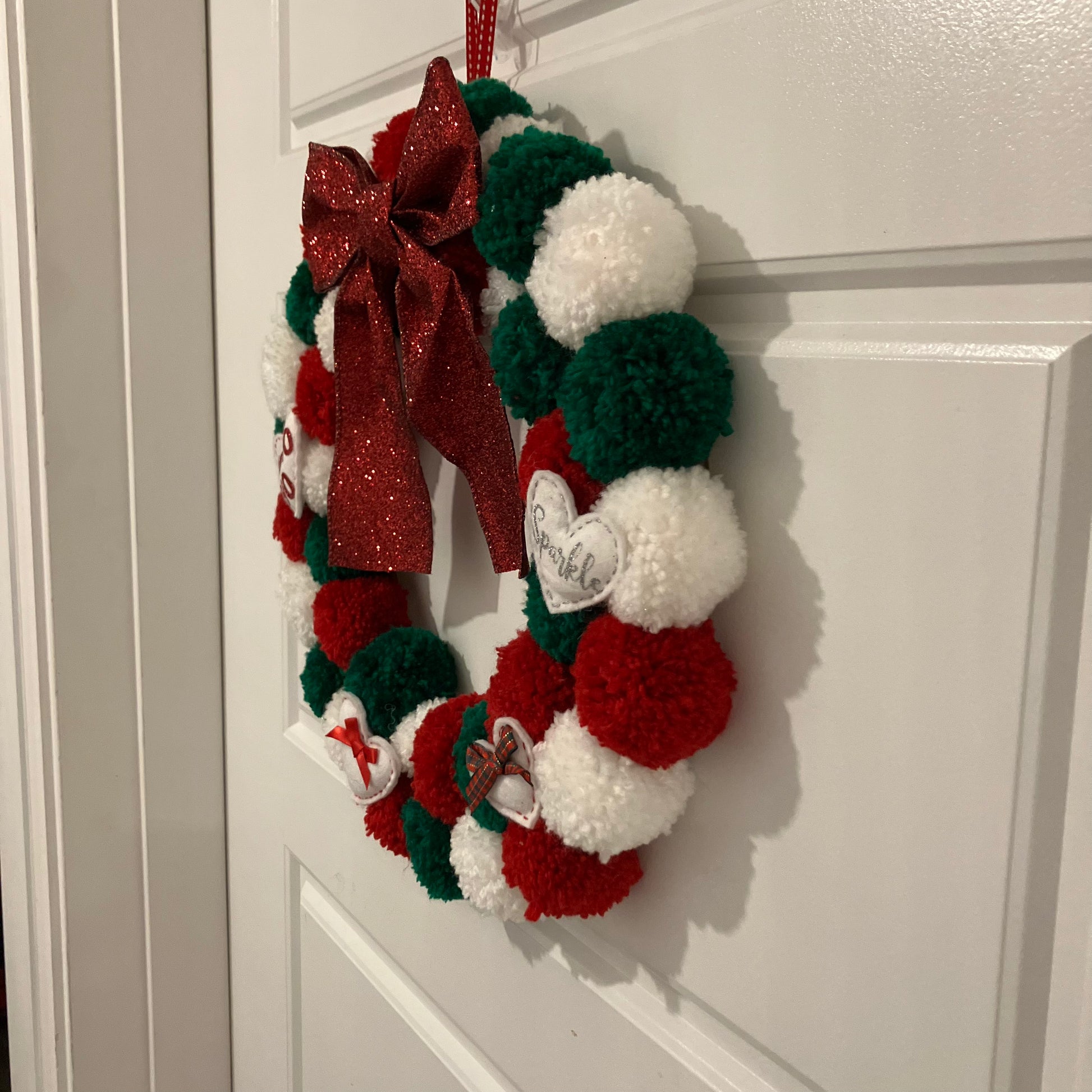 Handmade Christmas wreath with red, green, and white pom-poms, a red bow, and small white ornaments.
