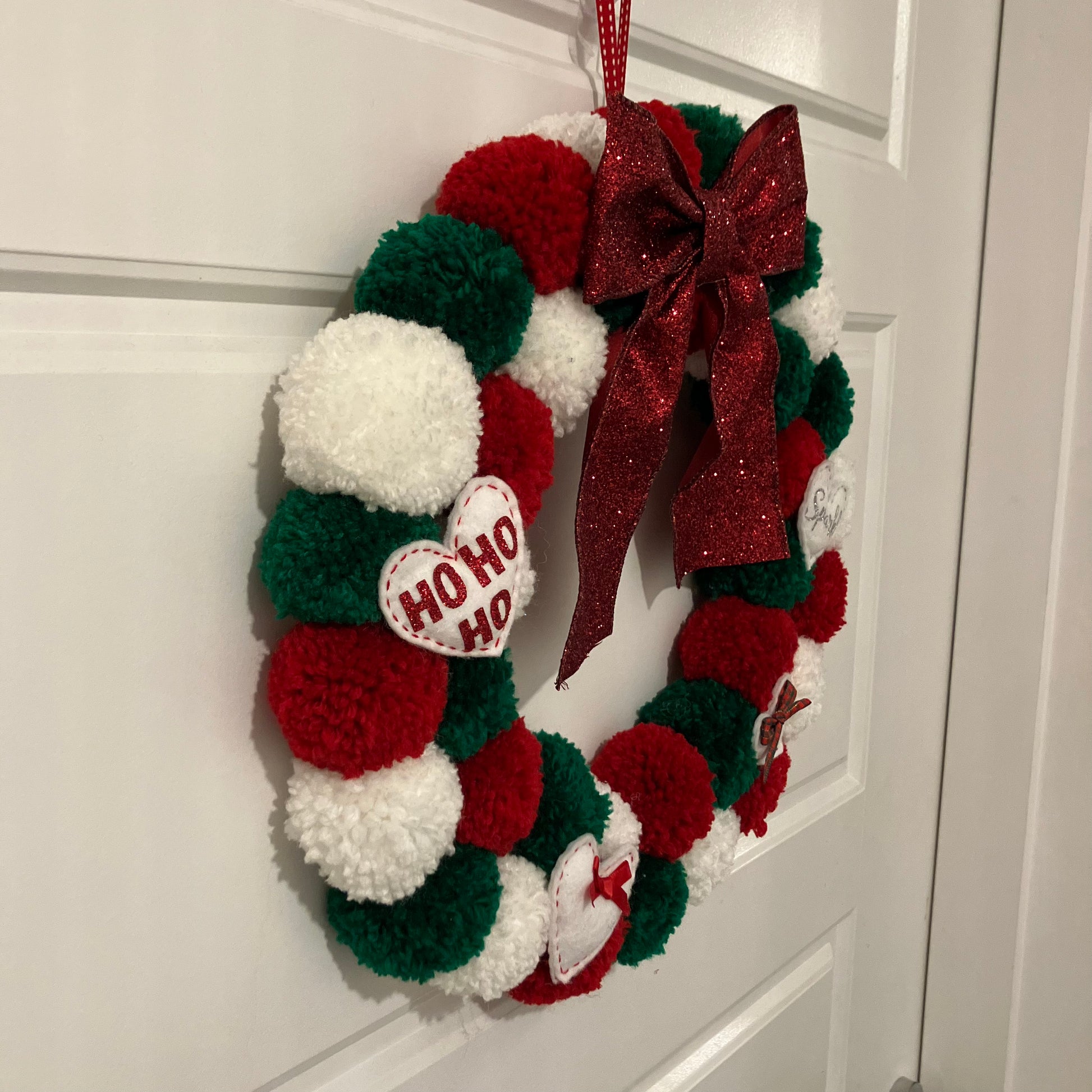 Handmade Christmas wreath with red, green, and white pom-poms, a red bow, and a "HO HO HO" heart.