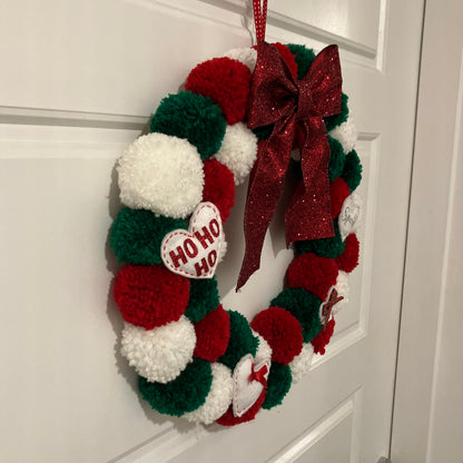 Handmade Christmas wreath with red, green, and white pom-poms, a red bow, and a "HO HO HO" heart.