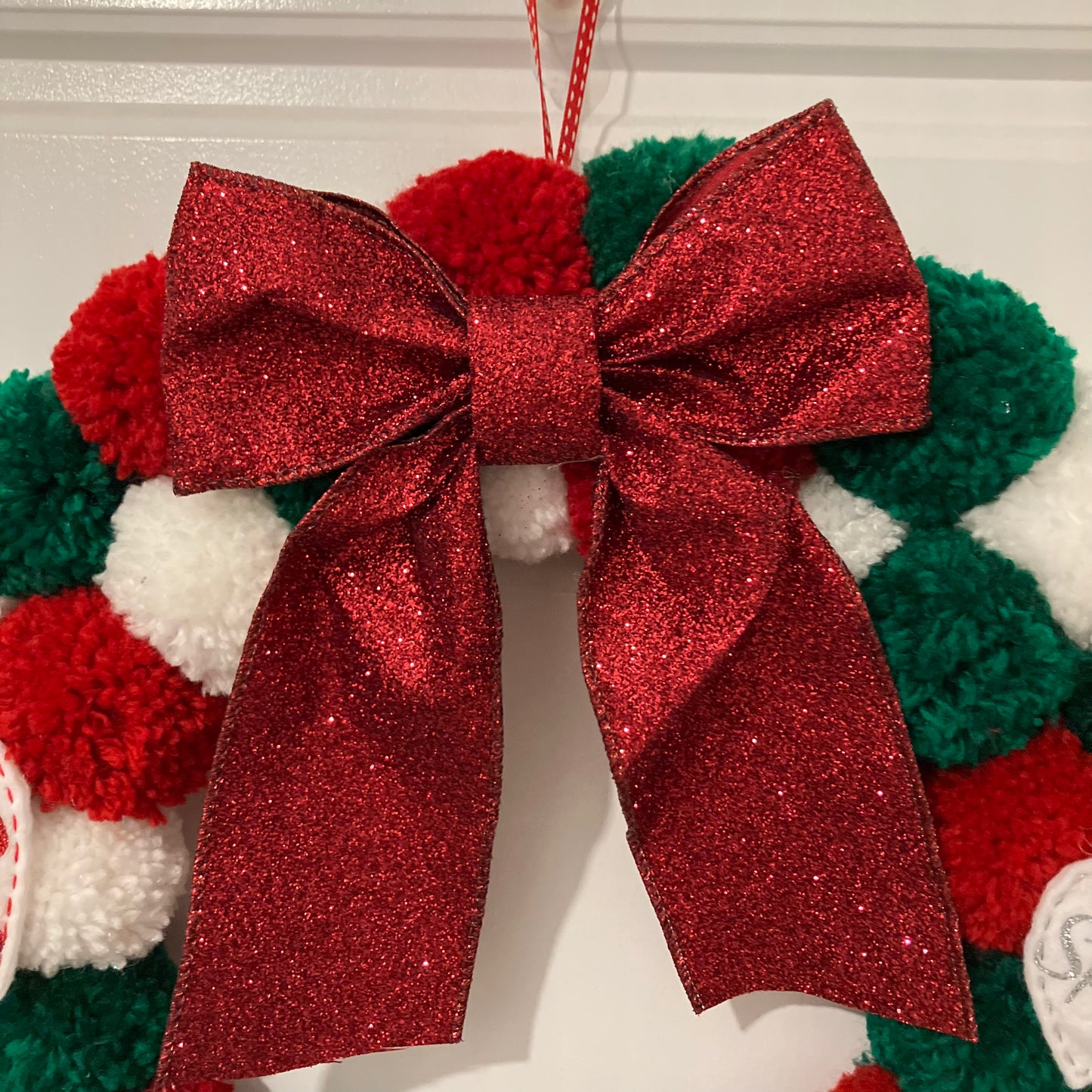 Red glitter bow on a Christmas wreath made of red, green, and white pom-poms.