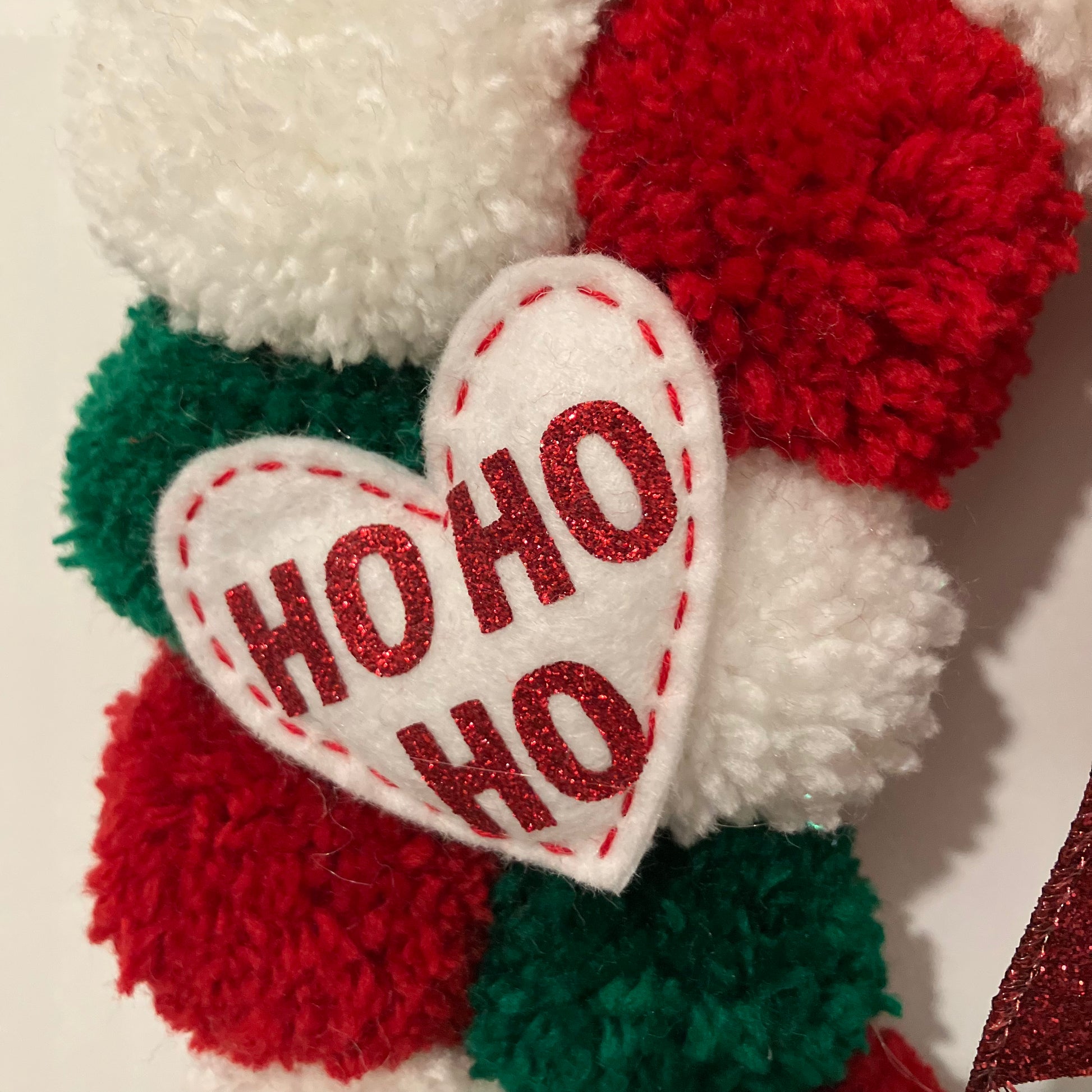 Close-up of a white felt heart with "HO HO HO" in red, on a red, white, and green pom-pom wreath.