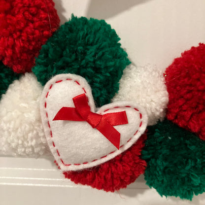 Close-up of a white felt heart with a red bow, surrounded by red, green, and white pom-poms.