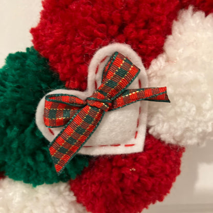Close-up of a white heart with a red and green plaid bow, on a wreath of red, green, and white pom-poms.