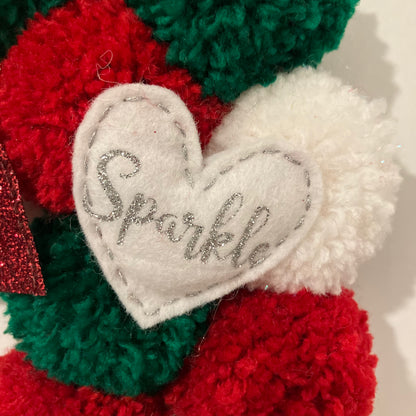 White felt heart ornament with "Sparkle" embroidered in silver, surrounded by red, green, and white pom-poms.