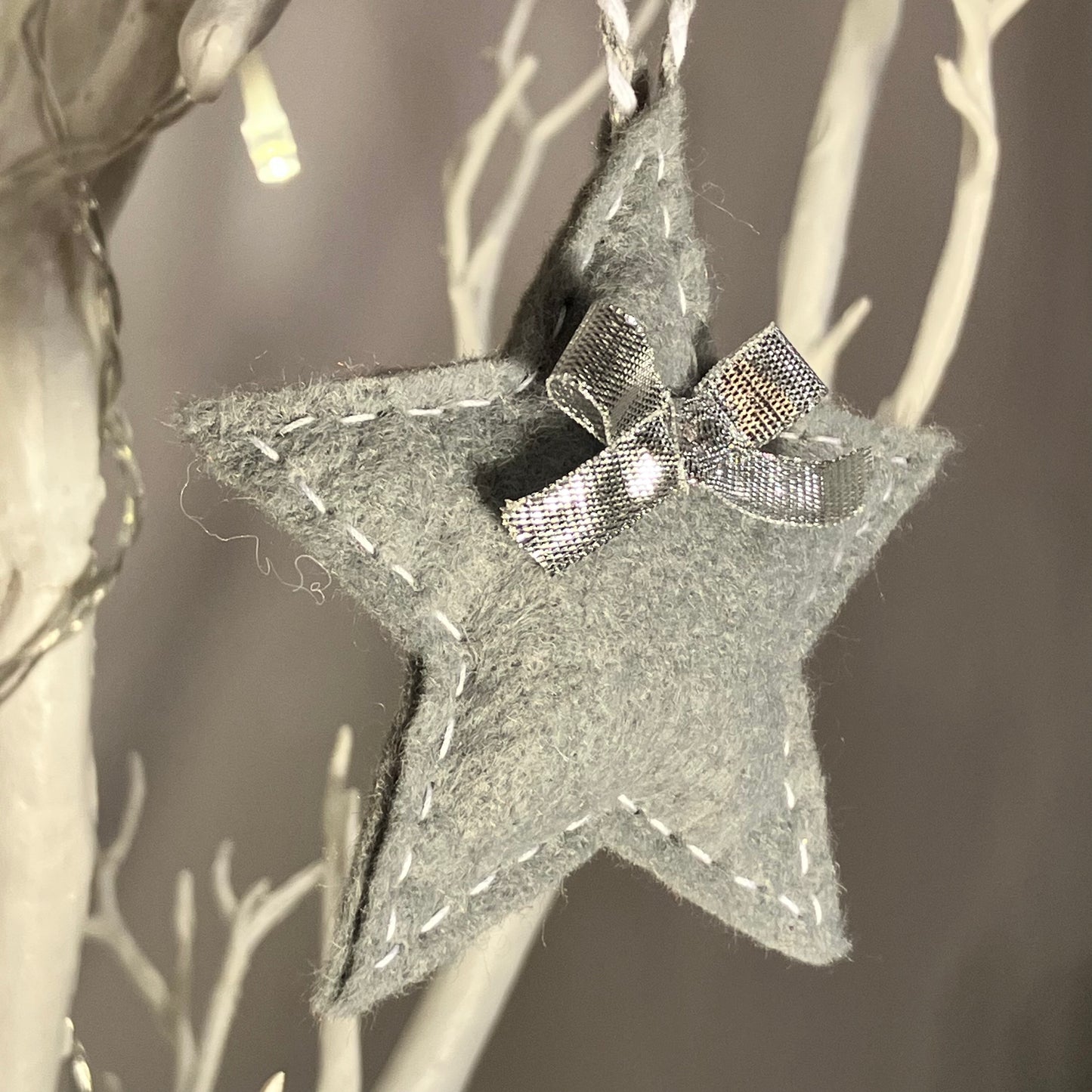 Grey felt star ornament with white stitching and a silver bow, hanging from a white branch.
