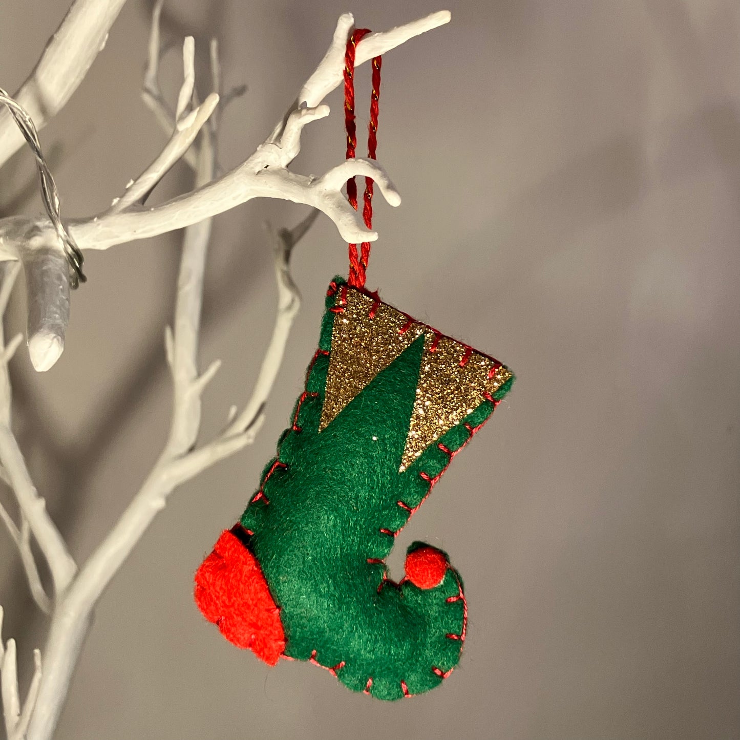 Green felt Christmas stocking with gold glitter cuff and red accents, hanging on a white branch.