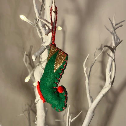 Green felt elf shoe ornament with gold glitter cuff and red trim, hanging on a white branch.