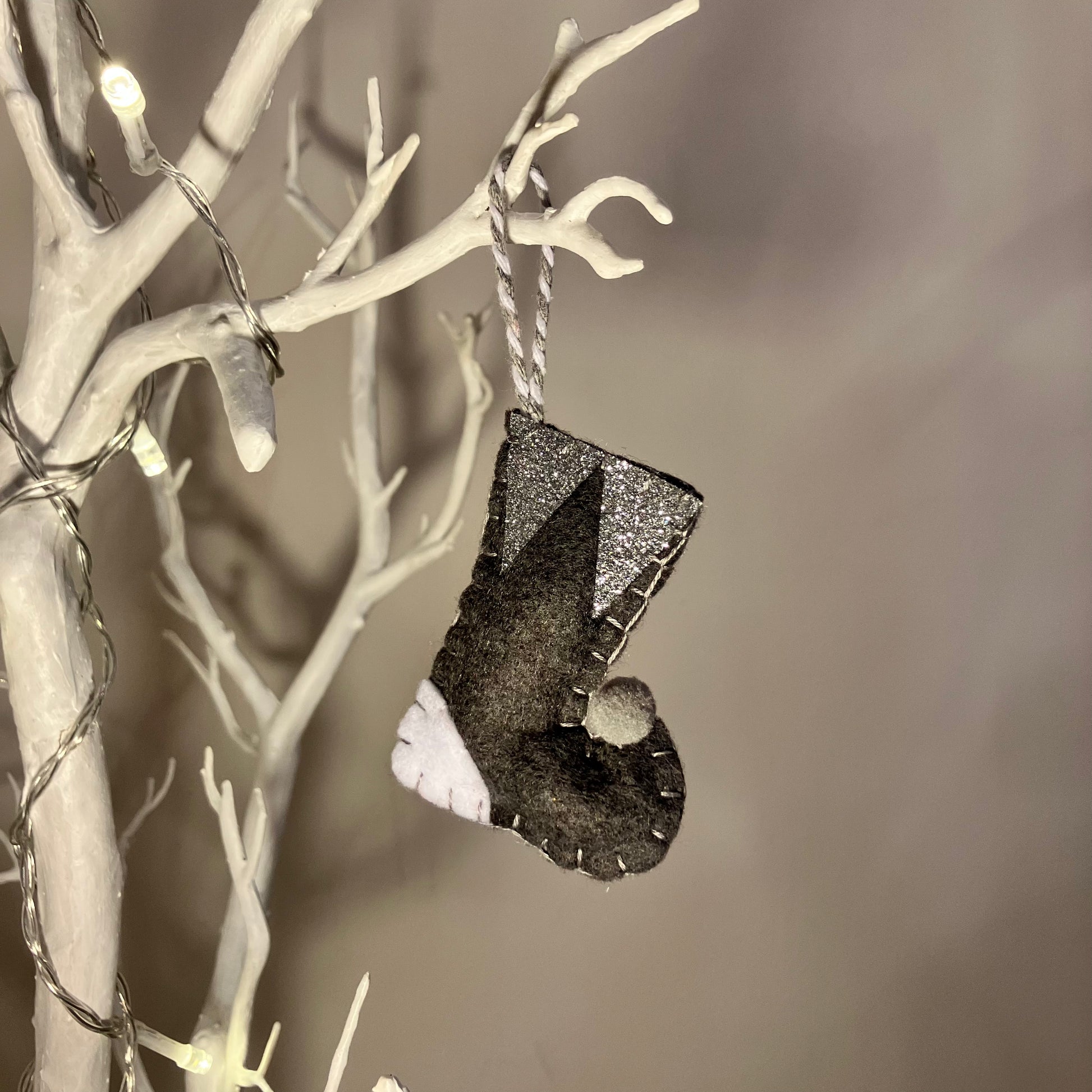 Handmade black felt Christmas stocking with white and silver accents hanging on a white branch.