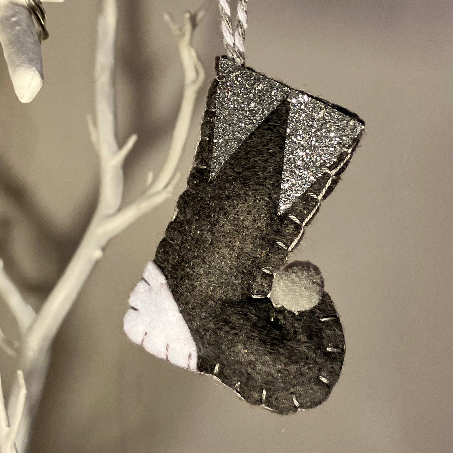 Handmade felt Christmas stocking ornament in black, white, and silver glitter, hanging from a white branch.