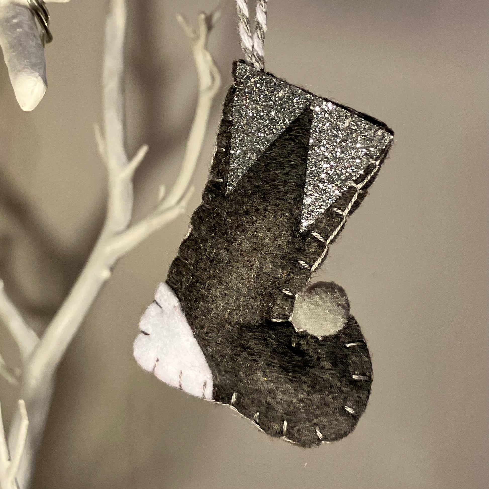Handmade felt Christmas stocking ornament in black, white, and silver glitter, hanging from a white branch.