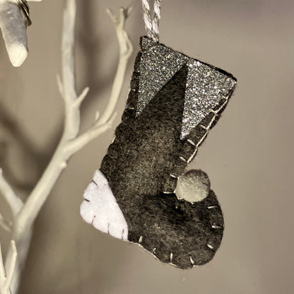 Handmade felt Christmas stocking ornament in black, white, and silver glitter, hanging from a white branch.