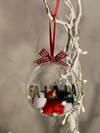Clear Christmas bauble with "FA LA LA" in gold, filled with red, white, and green pom-poms.