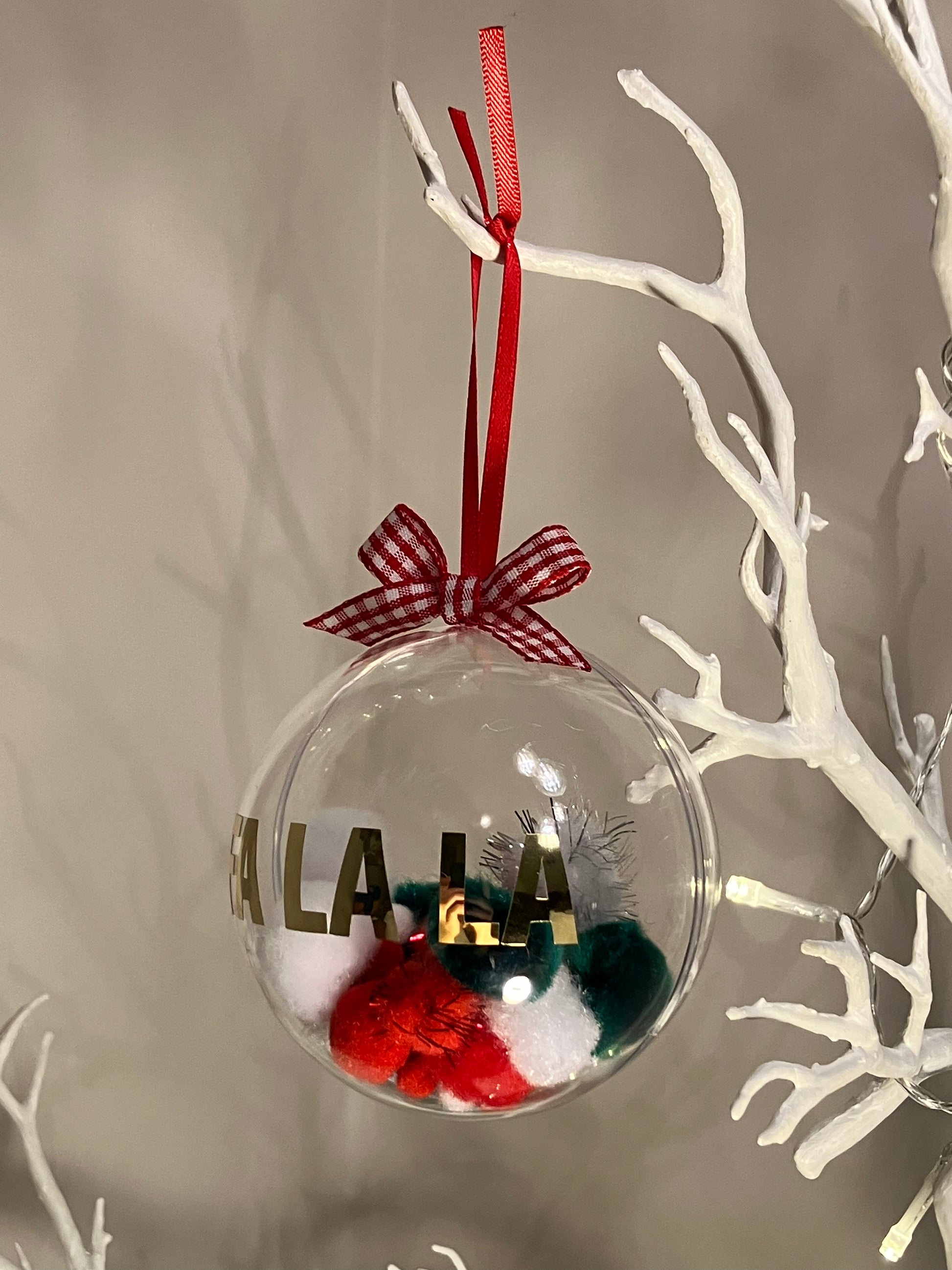 Clear ornament with red, green, white pom-poms and gold lettering, hanging from a white branch.