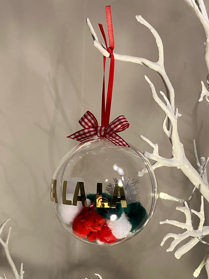 Clear ornament with red, green, white pom-poms and gold lettering, hanging from a white branch.