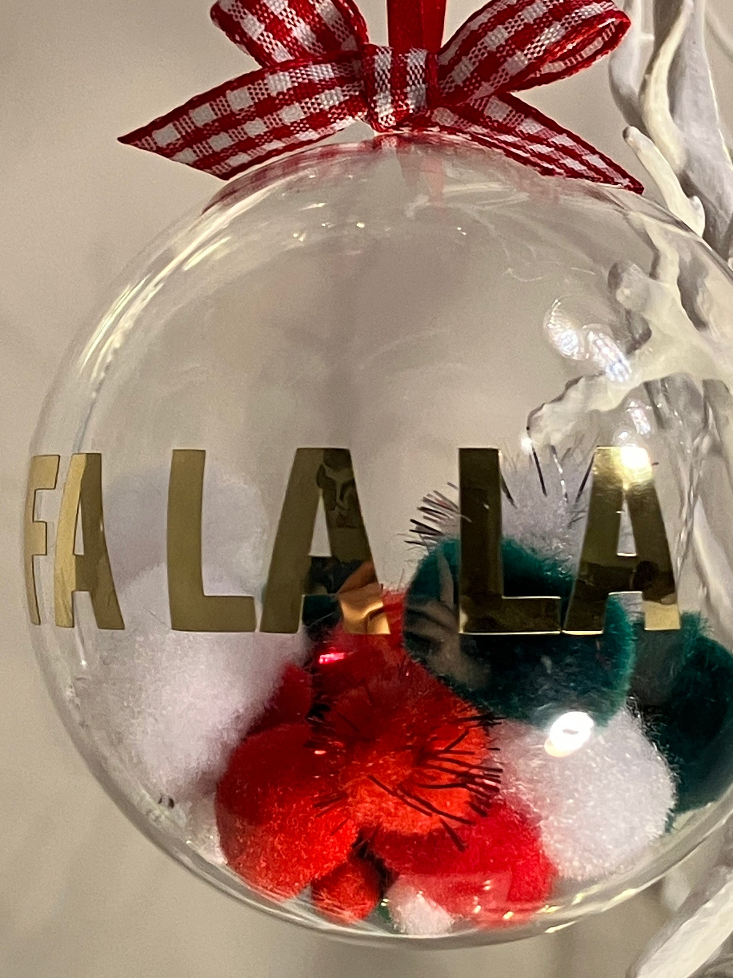 Clear Christmas ornament with "FA LA LA" in gold, filled with red, white, and green pom-poms.