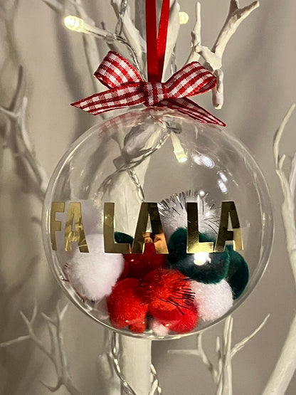Clear Christmas ornament with "FA LA LA" in gold, filled with red, white, and green pom-poms.