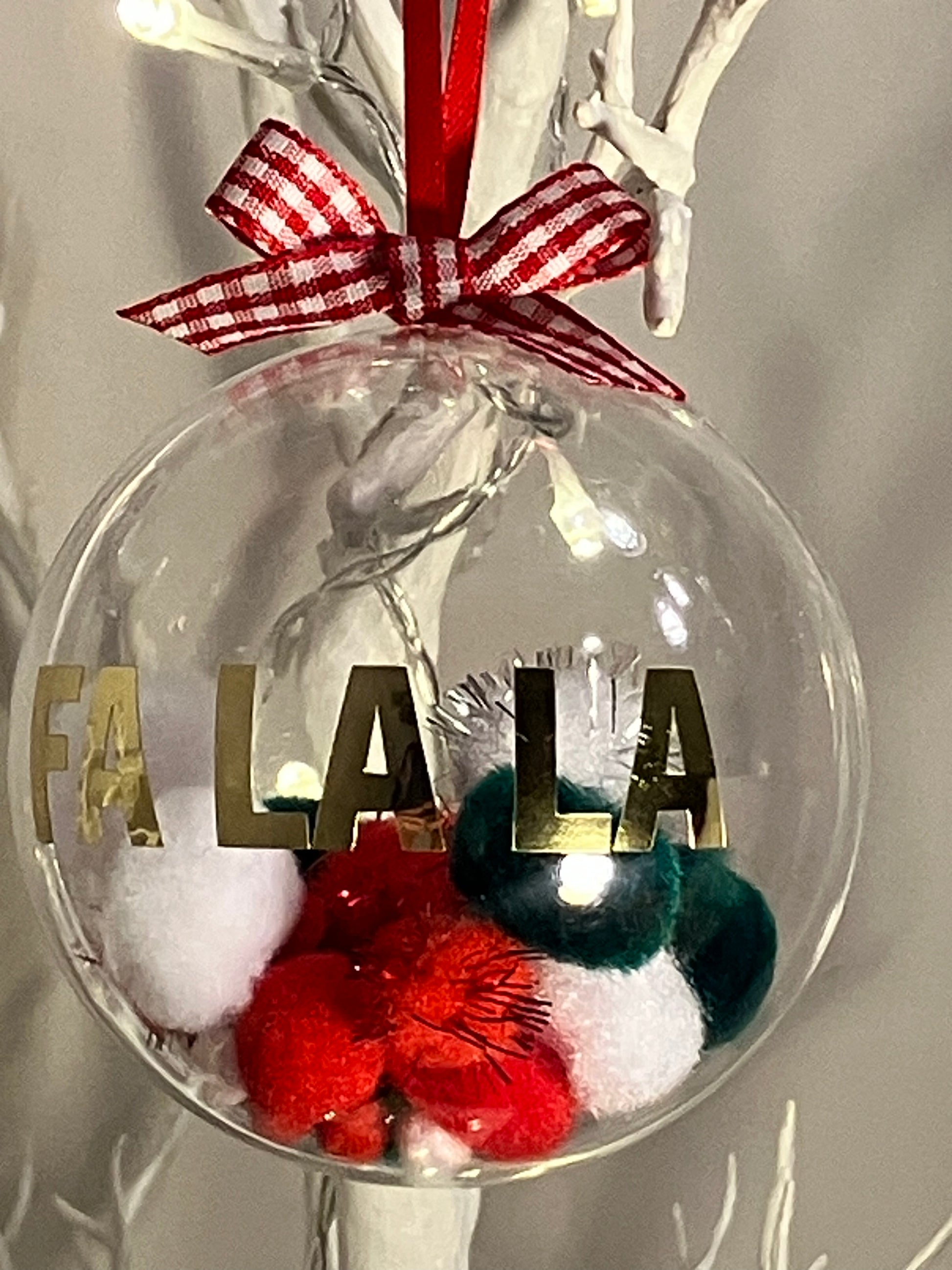 Clear ornament with "FA LA LA" in gold, filled with red, white, and green pom-poms.