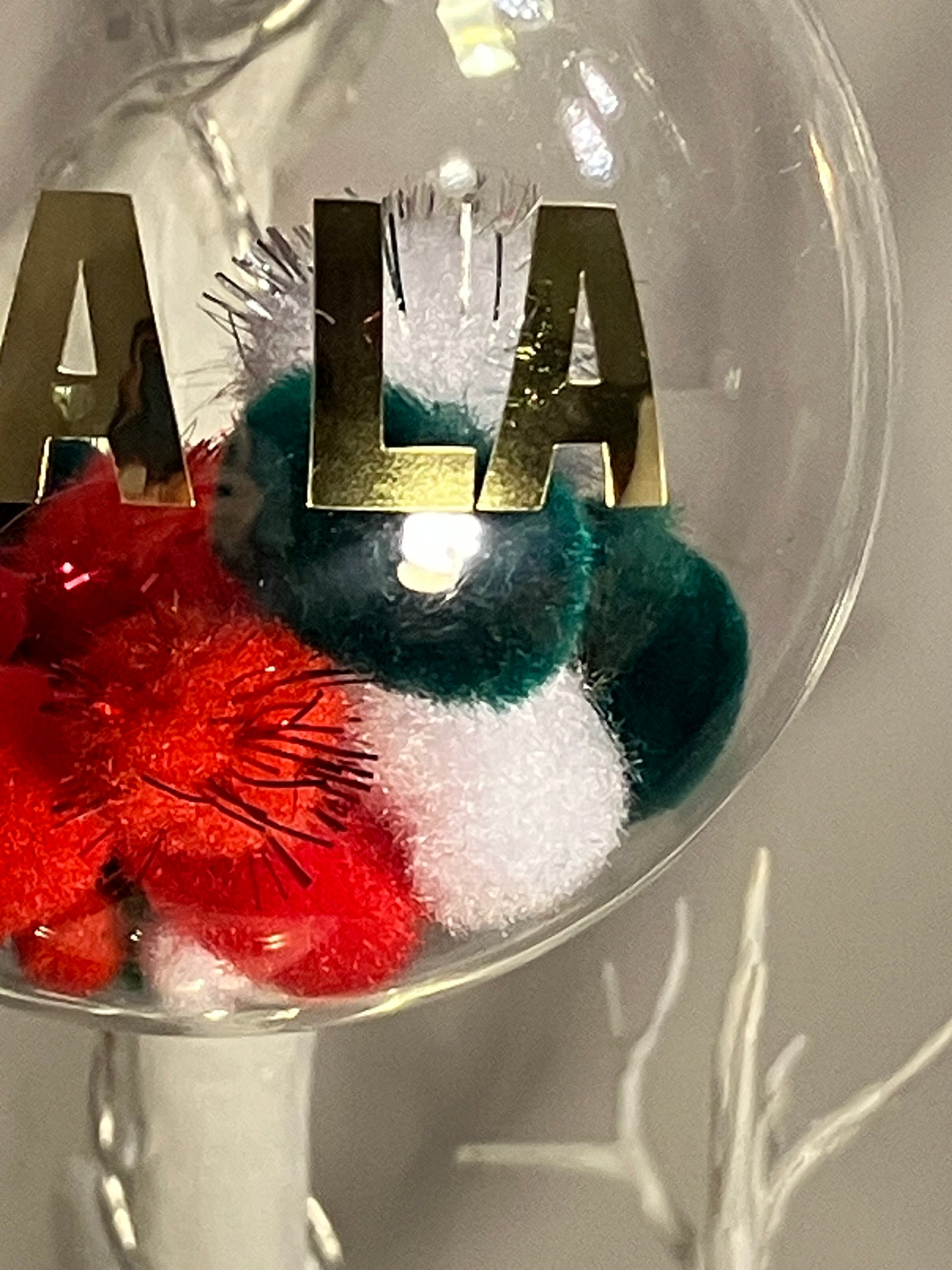 Clear ornament with gold letters "ALA" and red, green, and white pom-poms inside.