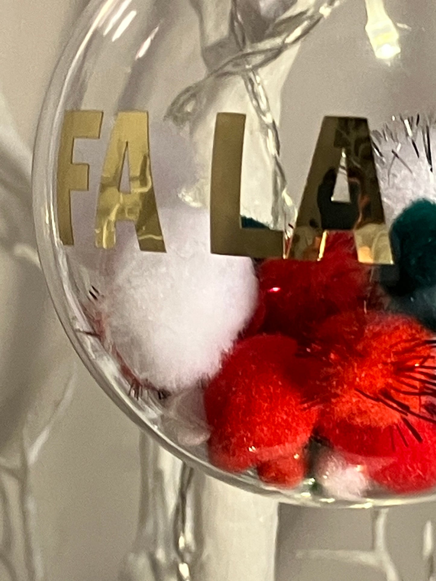 Clear ornament with gold "FA LA LA" text and red, white, and green pom-poms inside.