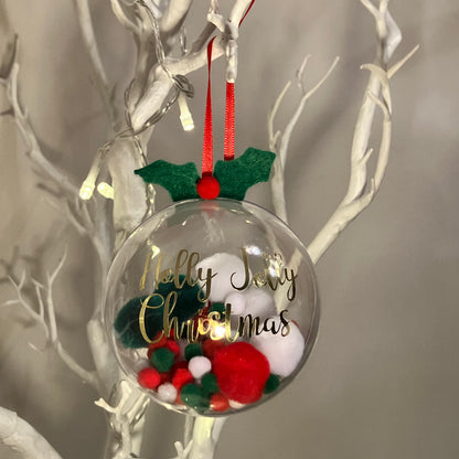 Clear Christmas bauble with green and red pom-poms and gold lettering.