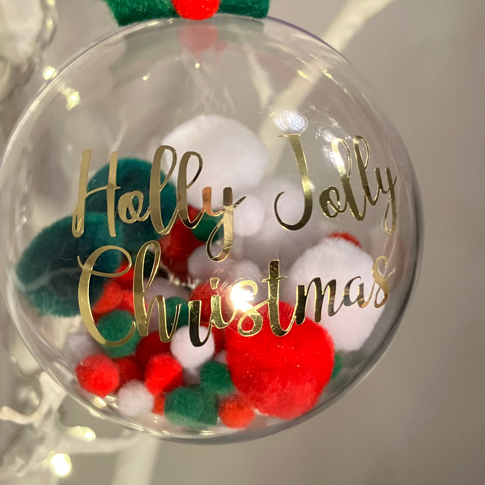 Clear Christmas bauble with red, white, and green pom-poms inside. Gold text reads "Holly Jolly Christmas".
