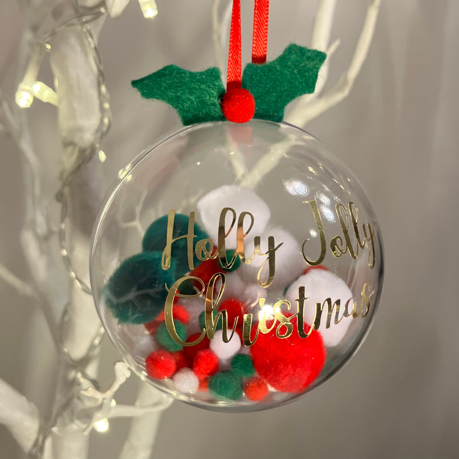 Clear Christmas bauble with "Holly Jolly Christmas" in gold, filled with red, green, and white pom-poms.