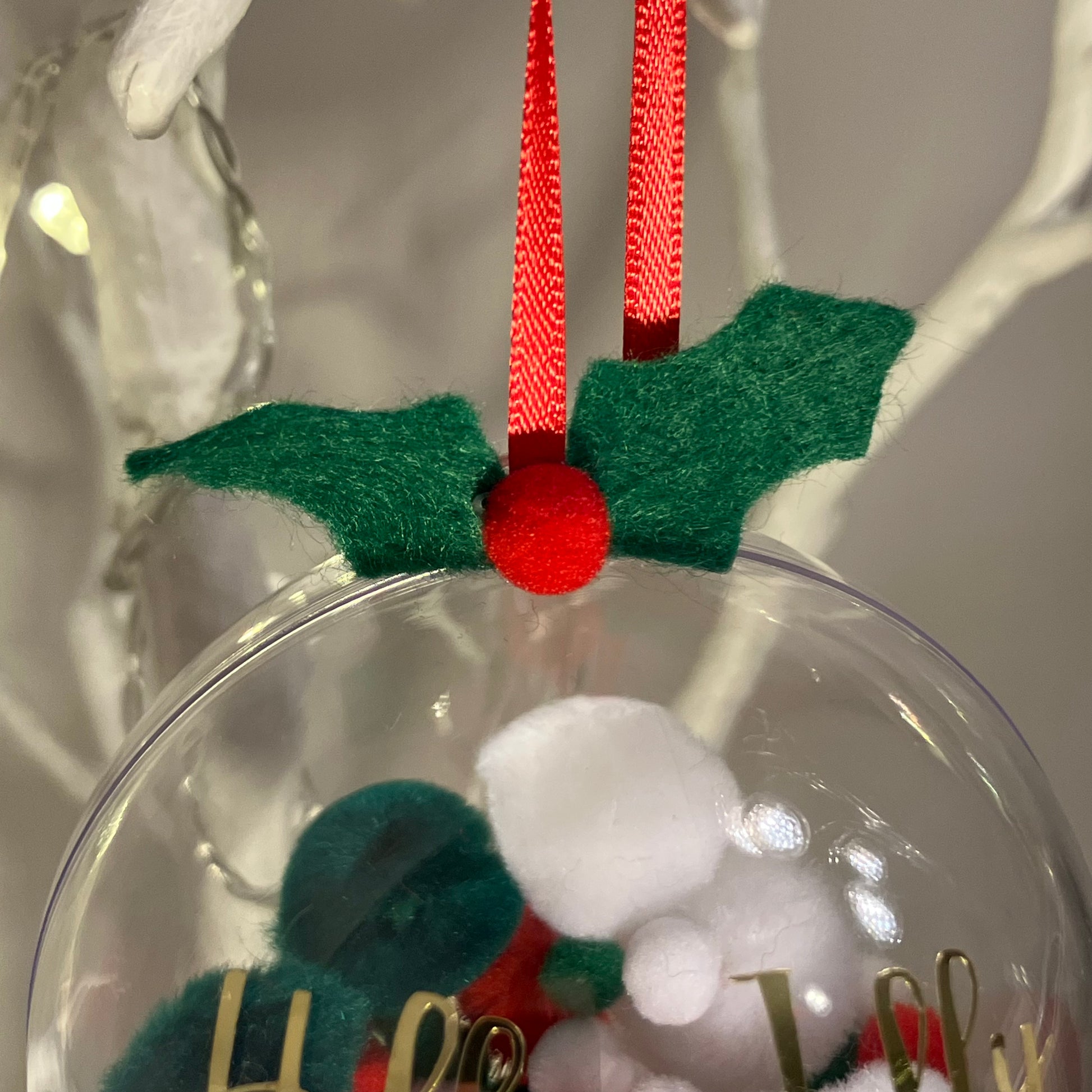 Clear Christmas bauble with red ribbon and green felt holly, containing two small figures.