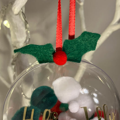 Clear Christmas bauble with red ribbon and green felt holly, containing two small figures.