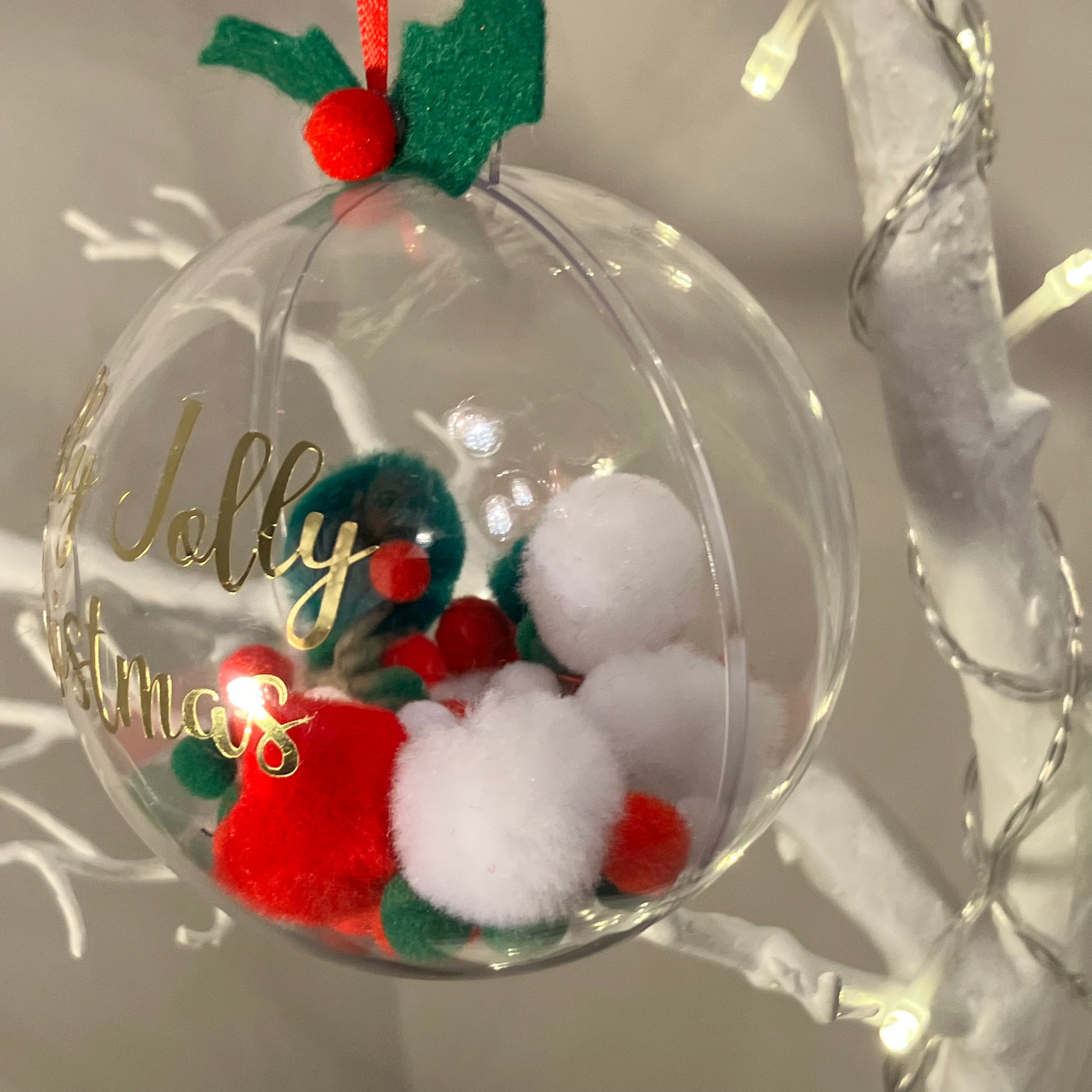 Clear Christmas bauble with "Jolly Christmas" text, filled with red, white, and green pom-poms.