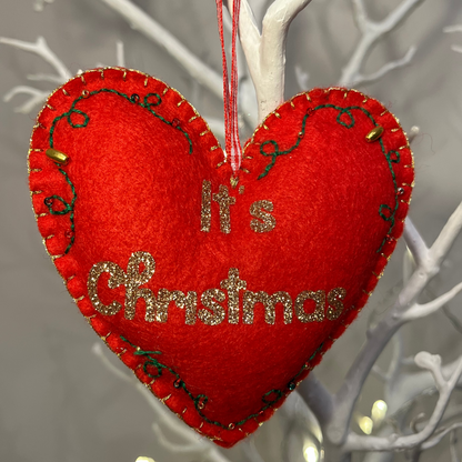 A red heart-shaped ornament with "It's Christmas" in gold glitter, hanging on a white branch.
