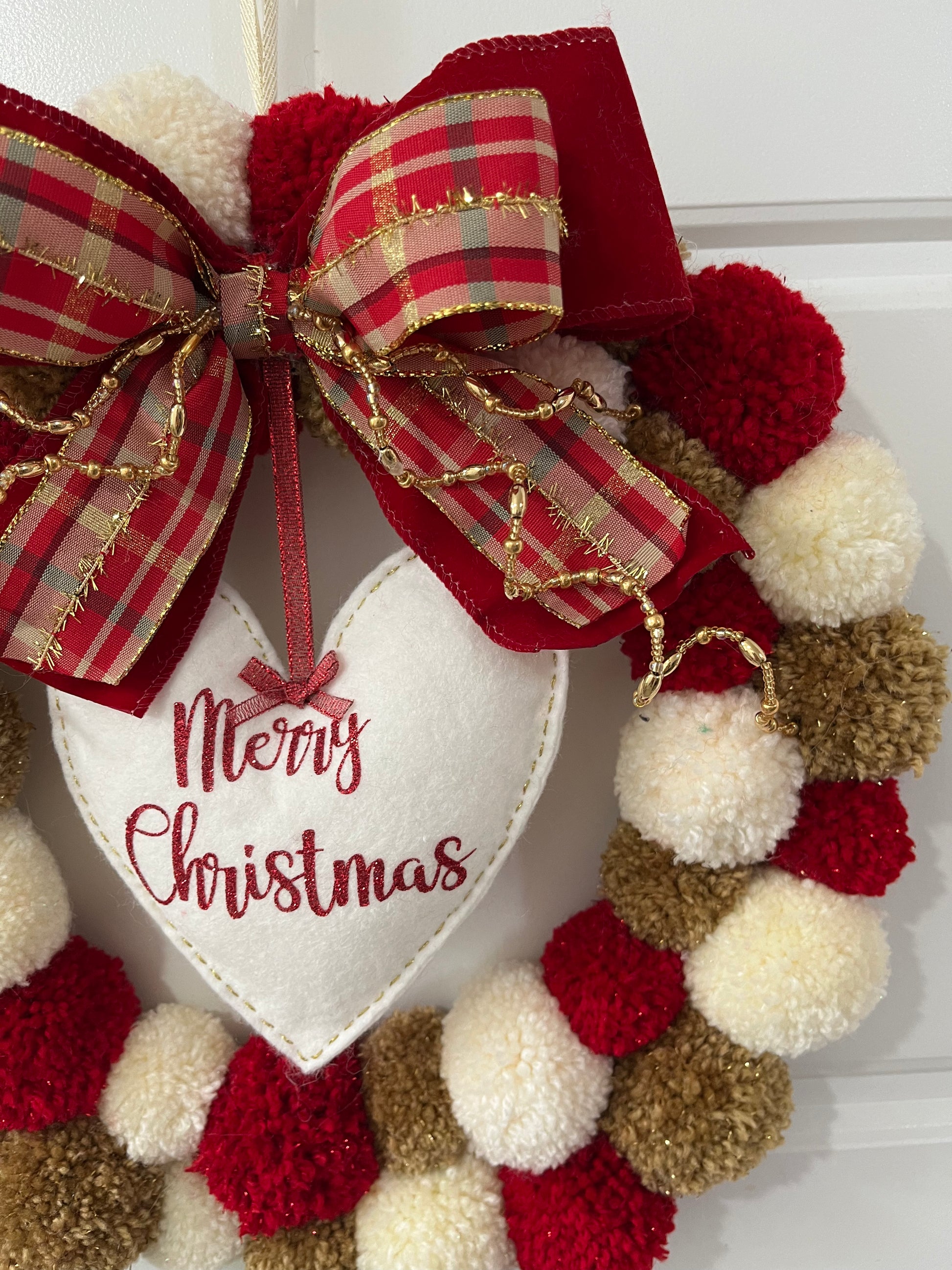 A close-up of a colorful pom-pom wreath adorned with a bow and "Merry Christmas" heart.