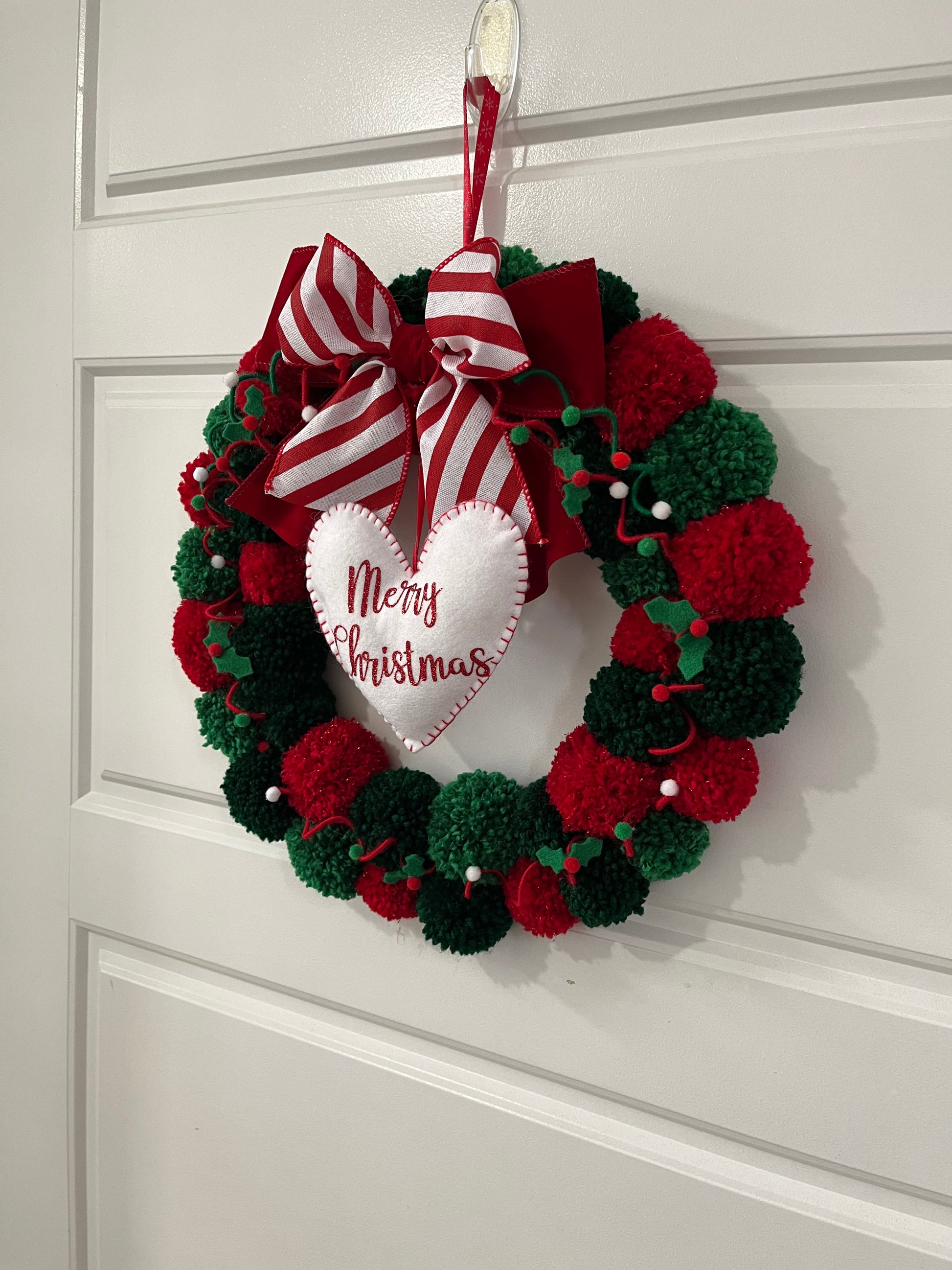 A red and green pom-pom wreath with a 'Merry Christmas' heart, hanging on a door.