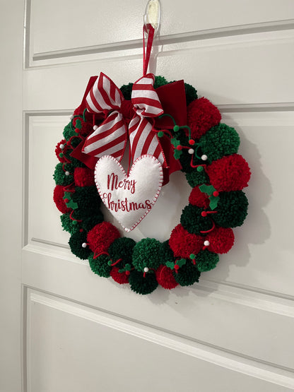 A red and green pom-pom wreath with a 'Merry Christmas' heart, hanging on a door.