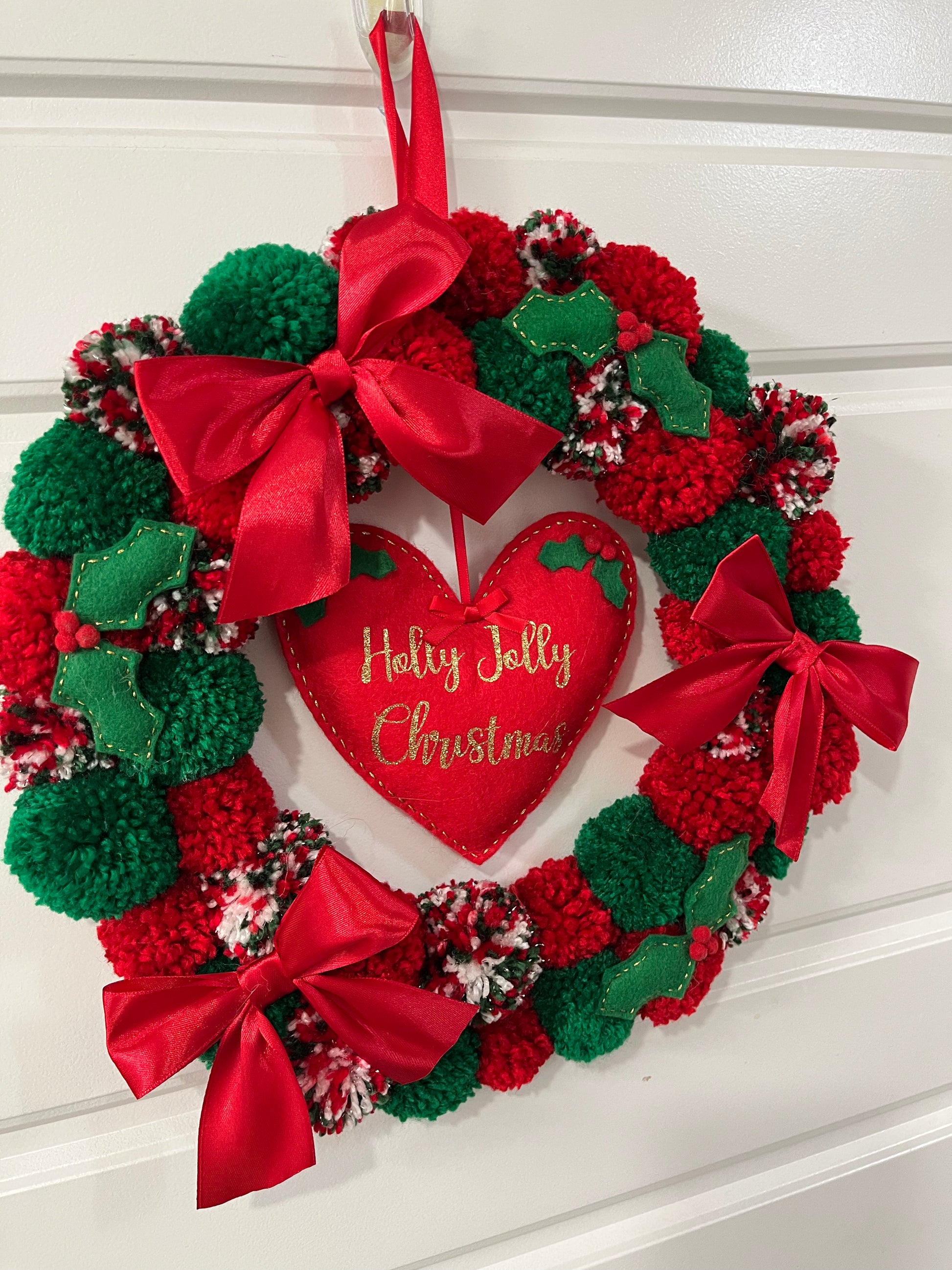 A handmade Christmas wreath with red and green pompoms, bows, and a 'Holly Jolly Christmas' heart.