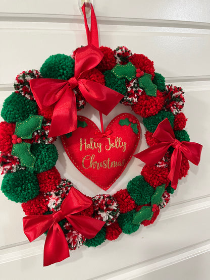 A handmade Christmas wreath with red and green pompoms, bows, and a 'Holly Jolly Christmas' heart.
